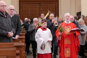 Der Pfarrer geht singend durch den Mittelgang der Kirche. Hinter ihm spielen Musikanten.