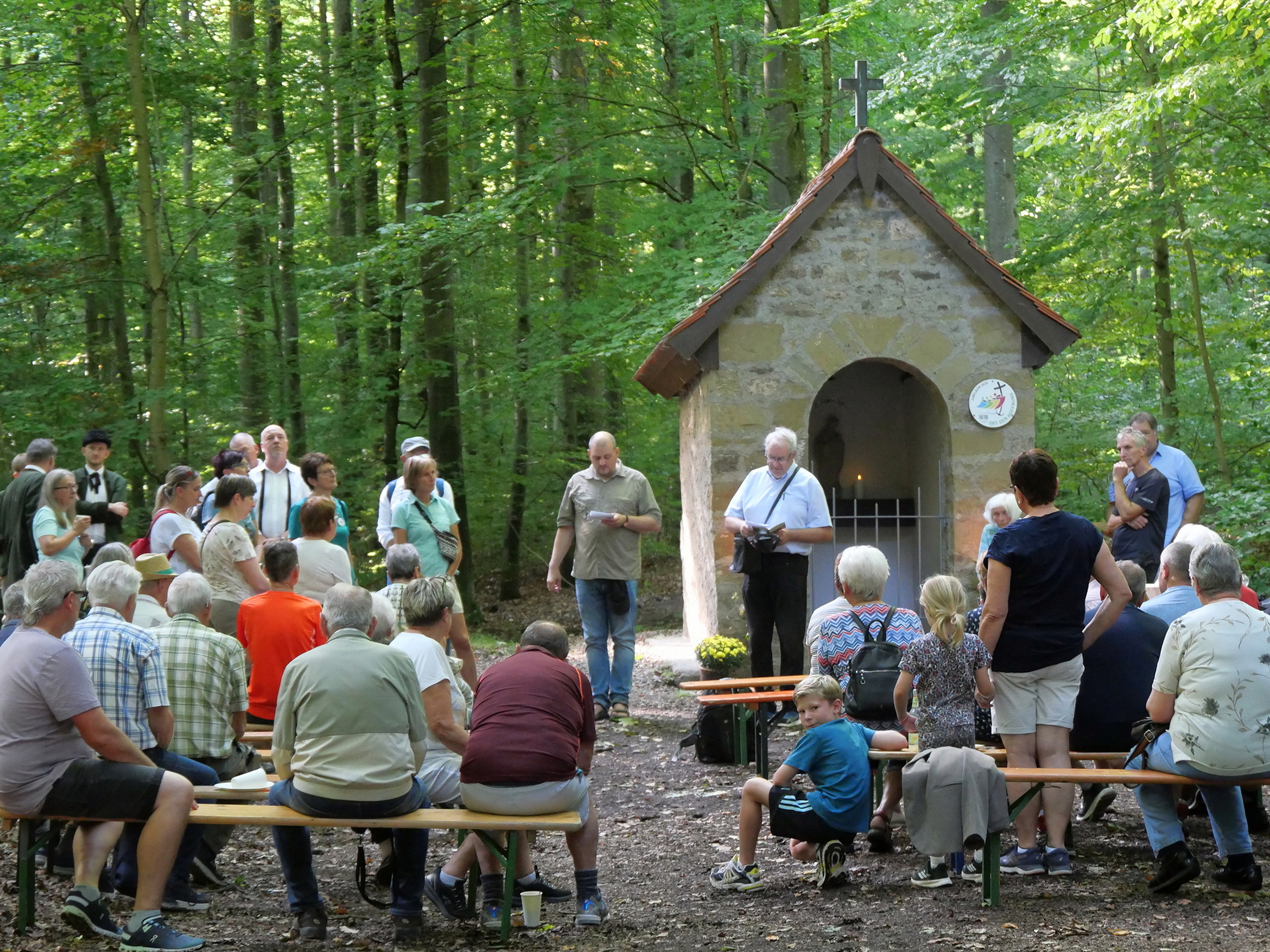 Blick zur kleinen Waldkapelle. Davor stehen und sitzen Menschen. An der Kapelle ist die Plaktette angebracht.