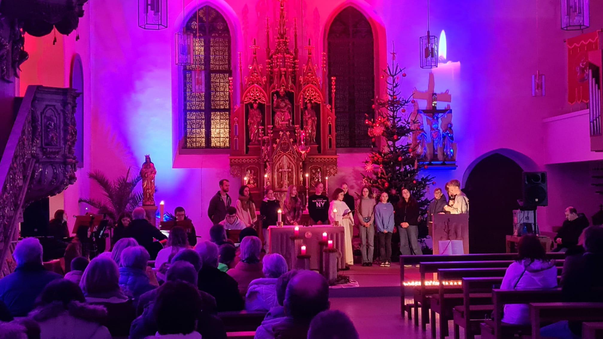 Blick auf den Altarraum der Kirche. Die Kirche ist mit Strahlern lila und rosa erleuchtet, im Altarraum stehen einige Jugendliche und lesen etwas vor.