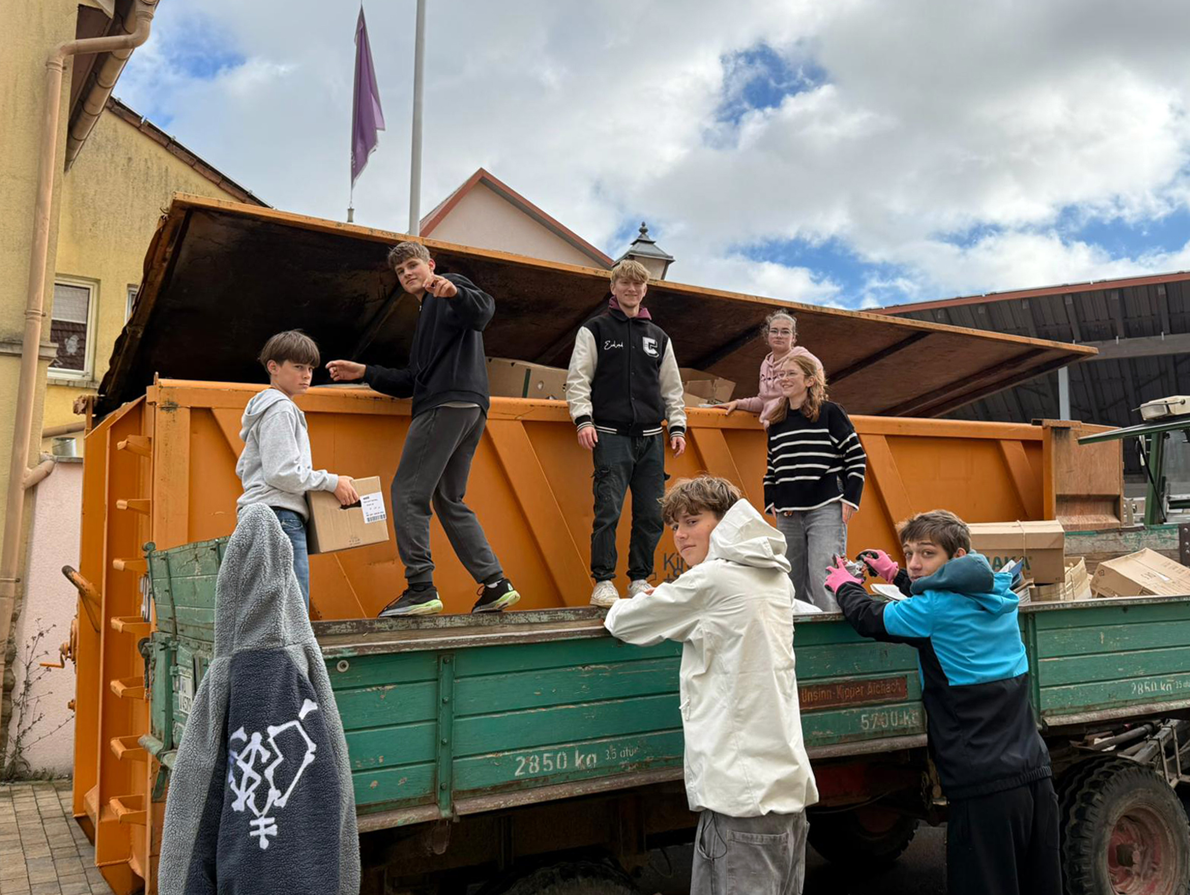 Jugendliche stehen um einen Altpapiercontainer und schauen in die Kamera.