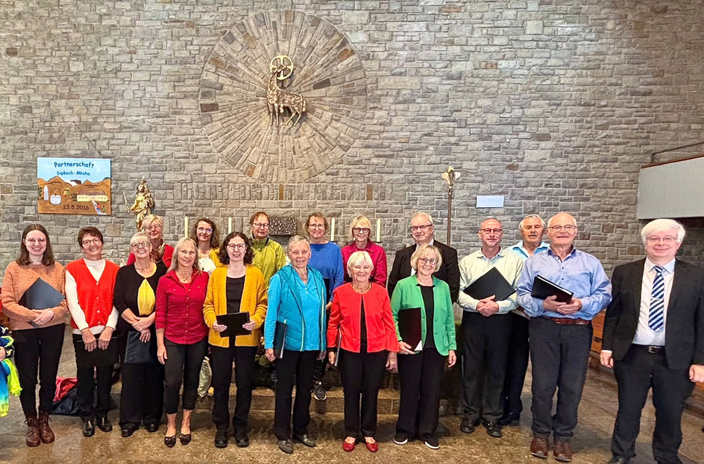 Das Bild ist ein Gruppenbild mit den Chormitgliedern und dem Chorleiter. Es ist in einer Kirche fotografiert worden. Alle freuen sich sichtlich.