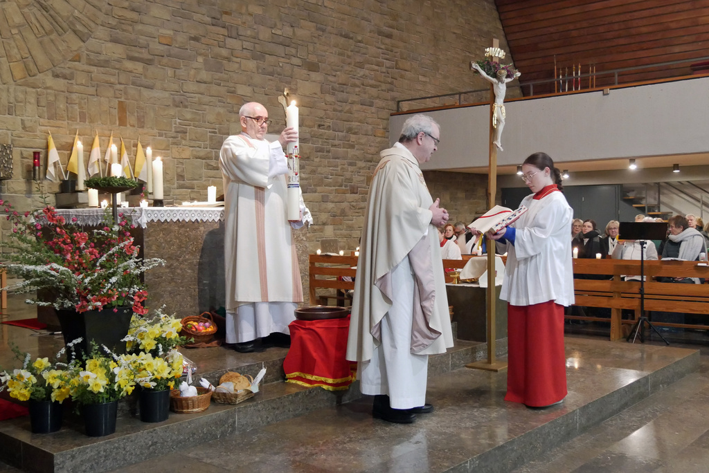 Der Diakon hält die brennende Osterkerze hoch. Vor ihm stehen der Pfarrer und eine Ministrantin.