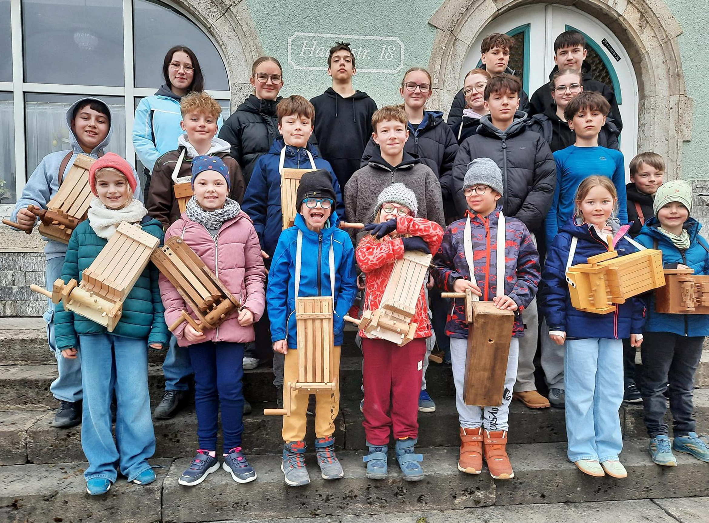 Das Bild zeigt eine Gruppe von etwa 20 Kindern und Jugendlichen, die vor einem Gebäude stehen. Einige Kinder halten hölzerne Geräuscheinstrumente. Alle sind warm angezogen, was auf kaltes Wetter hindeutet.
