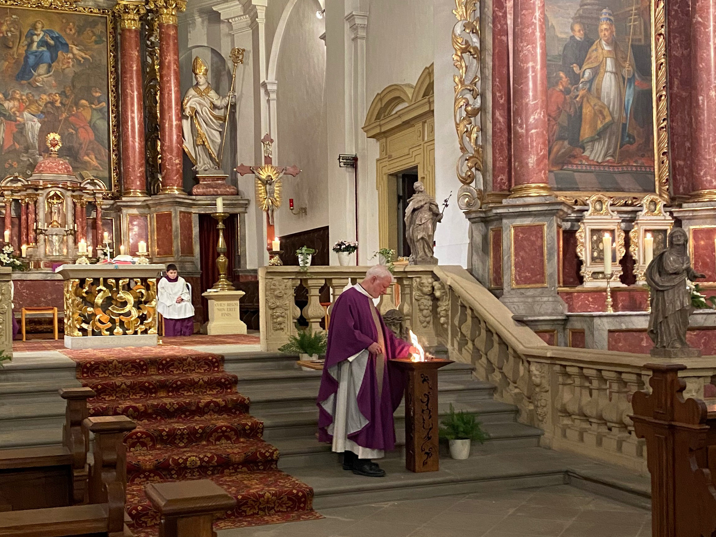 Ein Blick von den Kirchenbänken heraus nach vorne zum Altarraum. Auf den Altarstufen verbrennt Pater Matthäus Klein OSA die Palmzweige. Es ist ein kleines loderndes Feuer zu sehen.