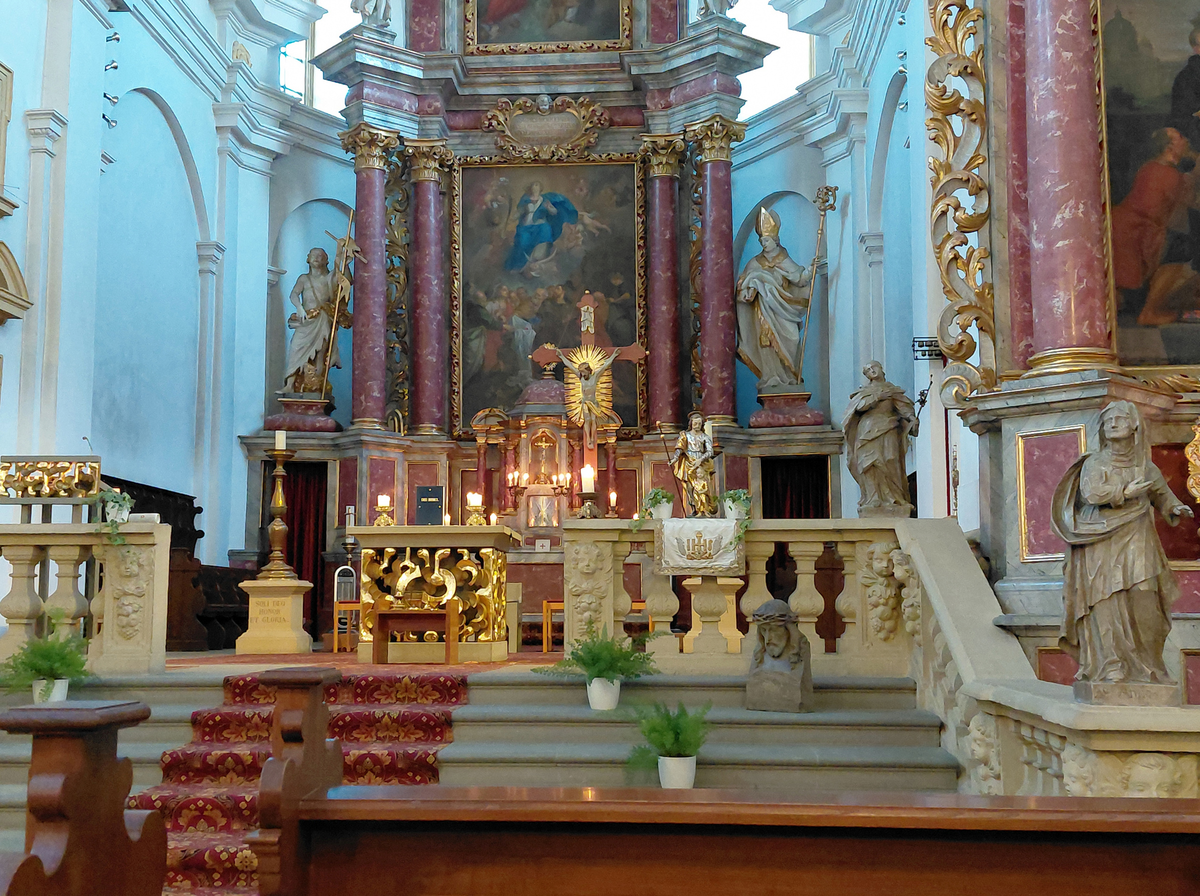 Das Bild zeigt einen prächtig dekorierten Altarbereich in einer Kirche. Es gibt mehrere Statuen von Heiligen, einen großen Altar mit einem Gemälde darüber, Kerzen und Blumenarrangements. Die Kirche hat hohe, gewölbte Decken und ist in hellen Farben gestrichen.