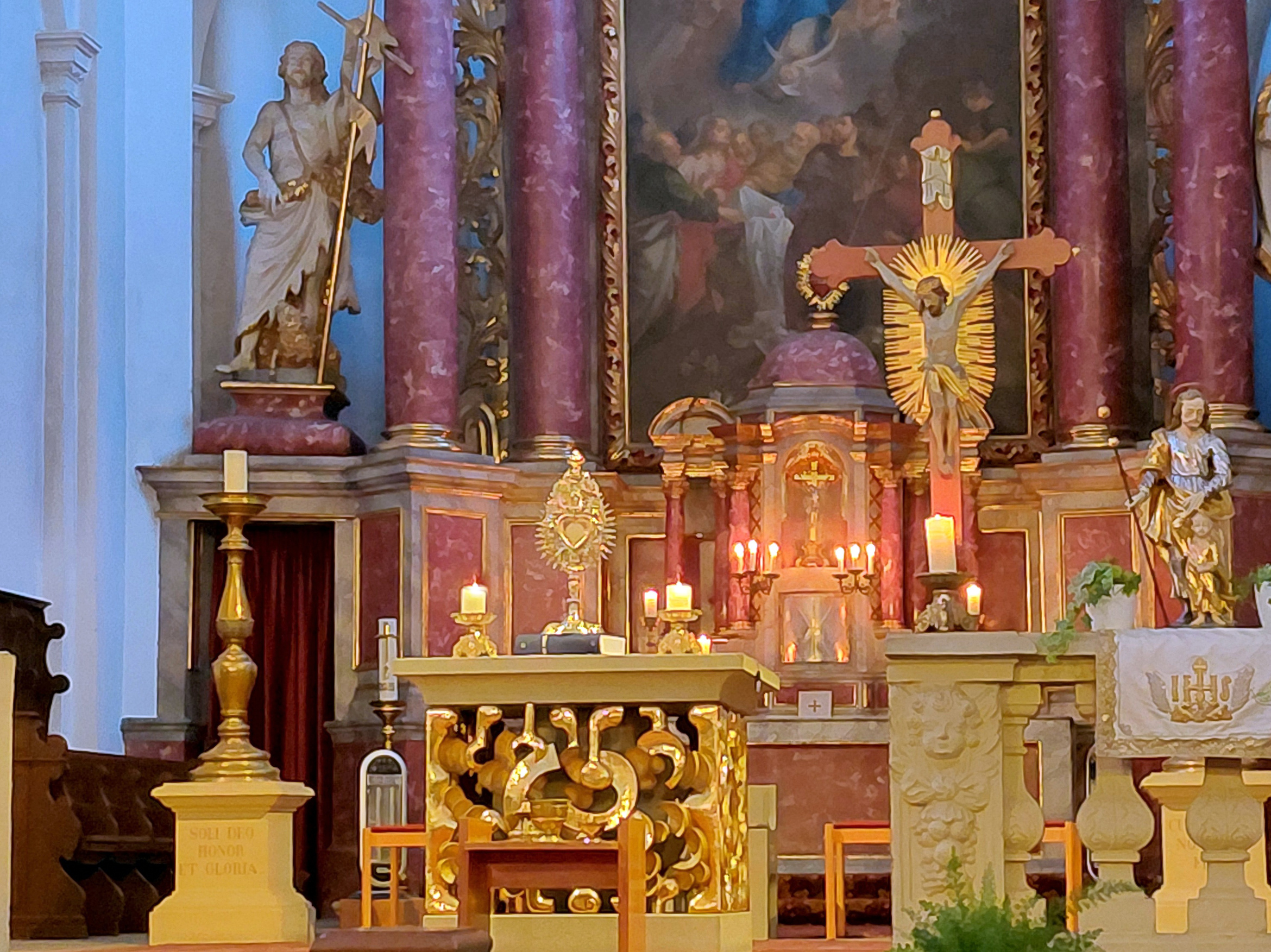 Das Bild zeigt den Altarbereich einer Kirche. Im Zentrum stehen ein vergoldeter Tabernakel und ein Kruzifix mit einer leuchtenden Aureole. Links und rechts befinden sich Heiligenstatuen, dahinter ein großes Gemälde und rosa marmorierte Säulen.