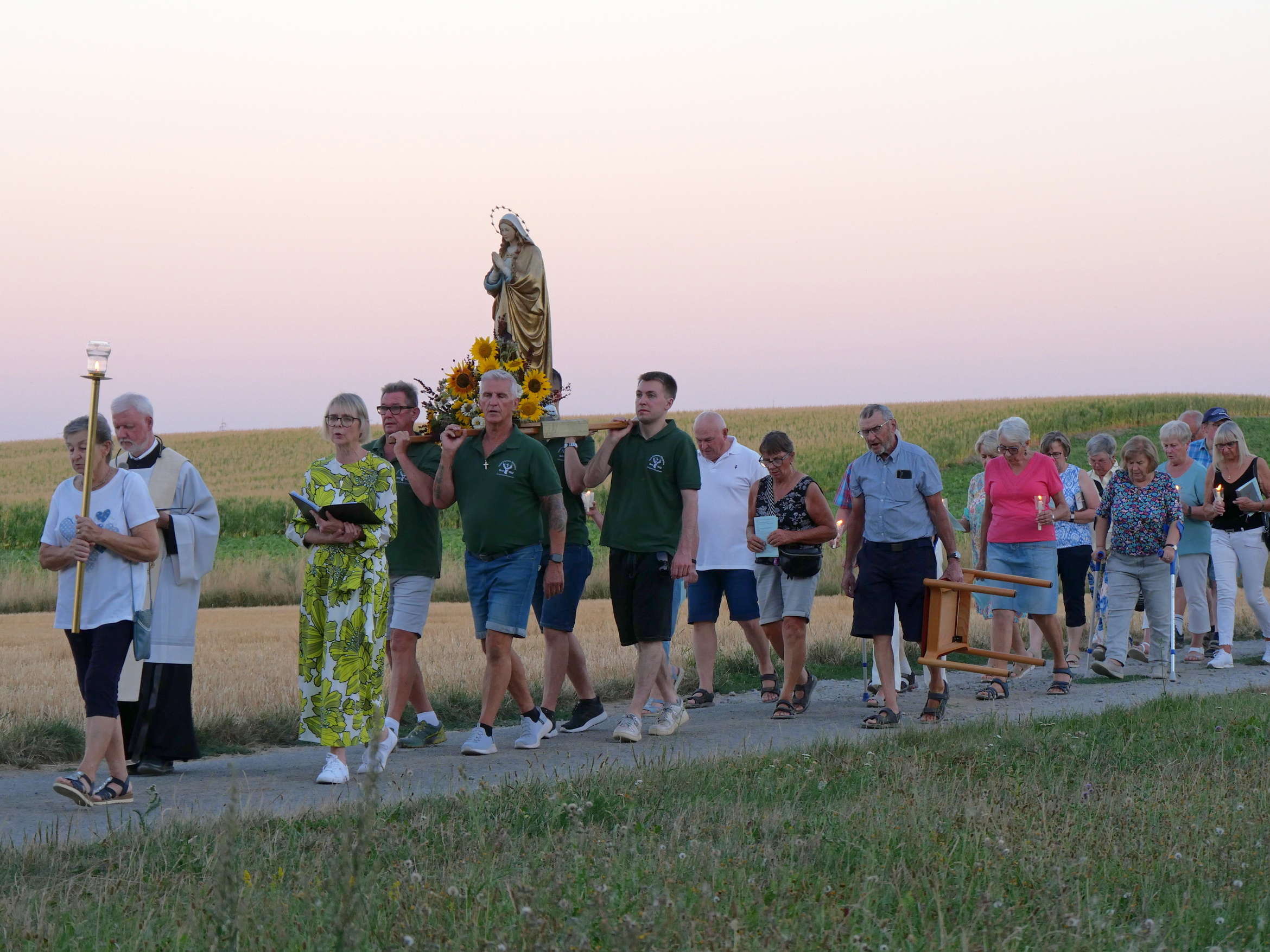 Das Bild zeigt mehrere Menschen bei einer Prozession. Im Hintergrund sind Felder. In der Mitte ist die Marienstatue.