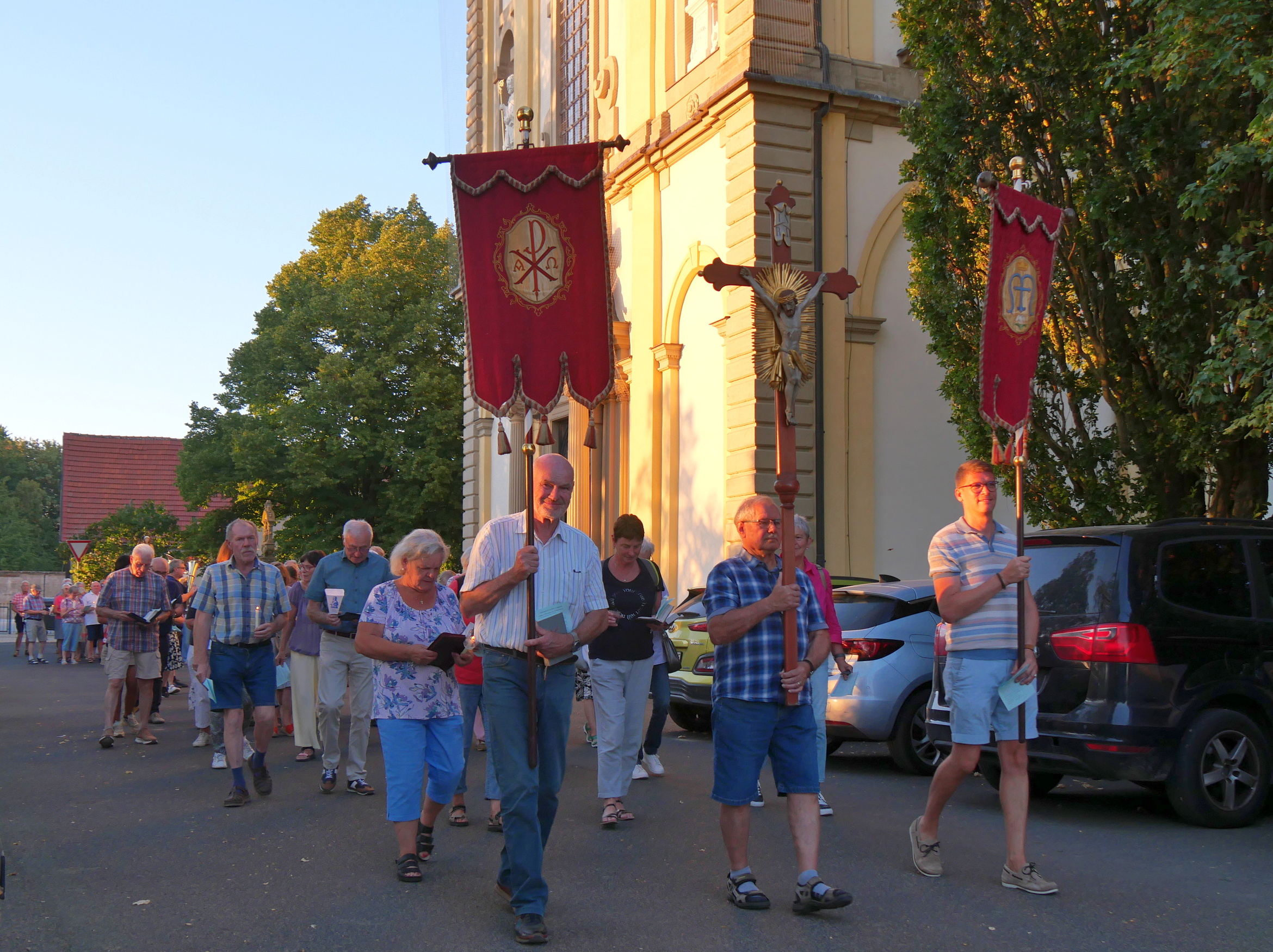 Das Bild zeigt eine Prozession mit Fahnen und einem Kreuz voraus. Im Hintergrund ist eine von der Sonne beleuchtete Kirche zu sehen.