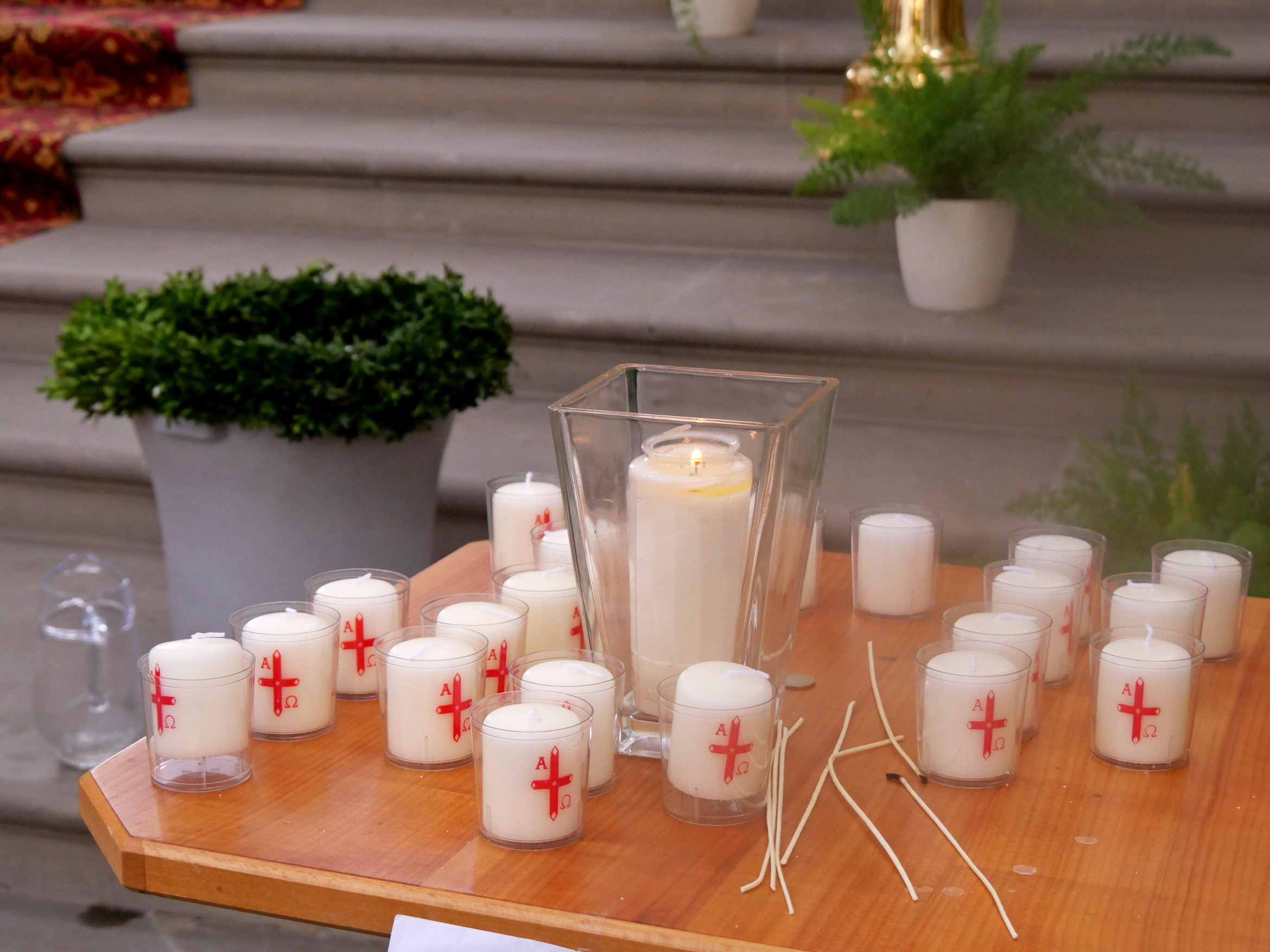 Das Bild zeigt einen Tisch mit mehreren weißen Kerzen in Gläsern, einige mit roten Kreuzen. Ein größeres Glas mit einer brennenden Kerze steht in der Mitte. Daneben befinden sich einige grüne Pflanzen in Töpfen.