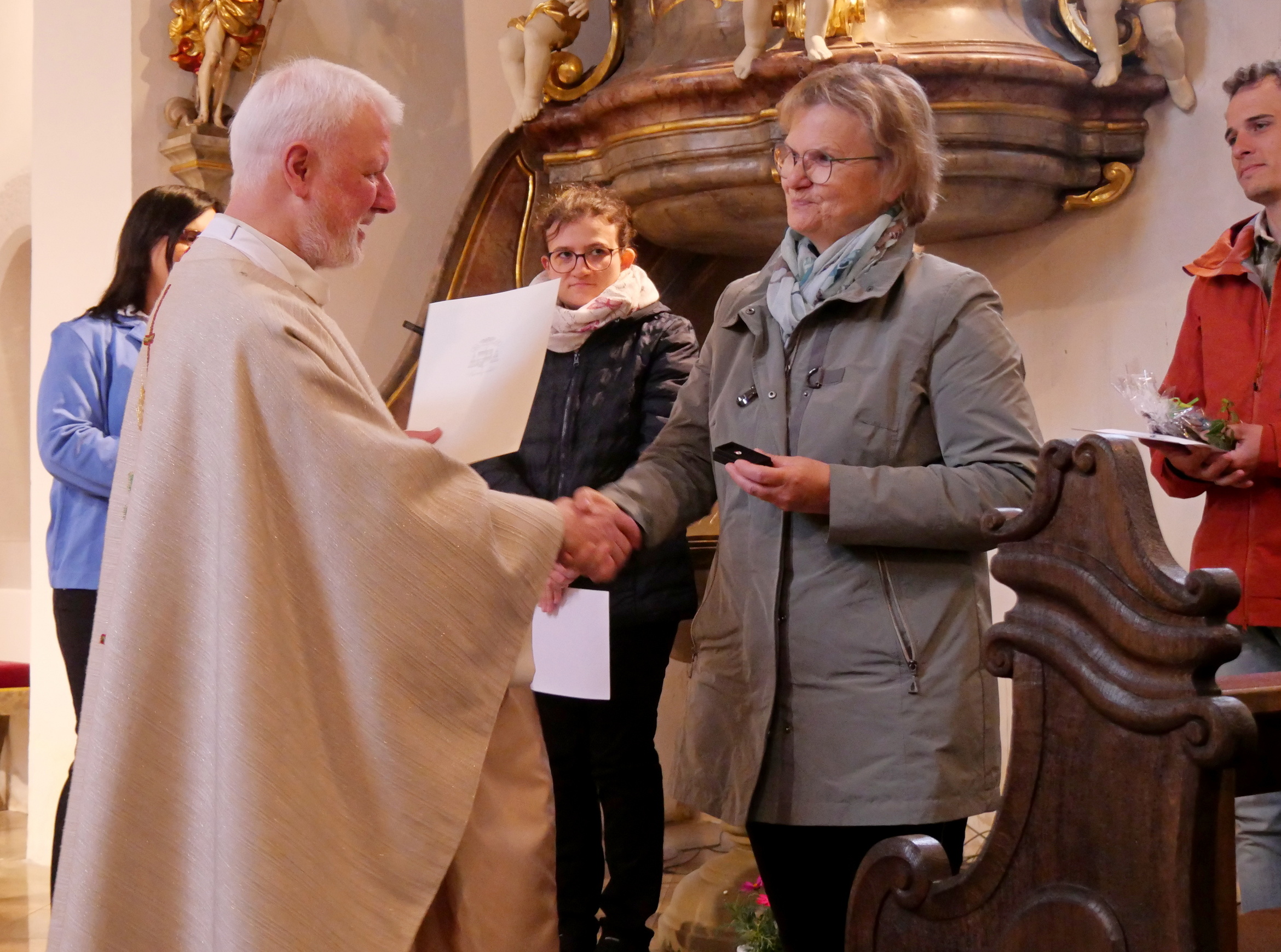 Das Bild zeigt einen Priester in einer hellen Robe, der einer Frau mit Brille und grauem Mantel die Hand schüttelt. Im Hintergrund stehen mehrere Personen, darunter eine mit blauer Jacke und eine mit rotem Mantel. Die Szene ist in einer Kirche.
