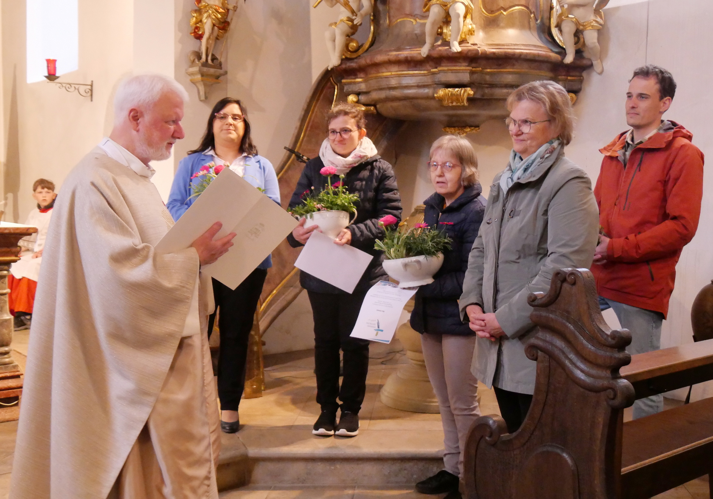 In einer Kirche steht ein Priester in einem weißen Gewand und spricht zu einer Gruppe von fünf Personen. Eine Frau hält einen Blumenstrauß und eine Urkunde. Im Hintergrund sind eine Kanzel und Kirchenbänke zu sehen.