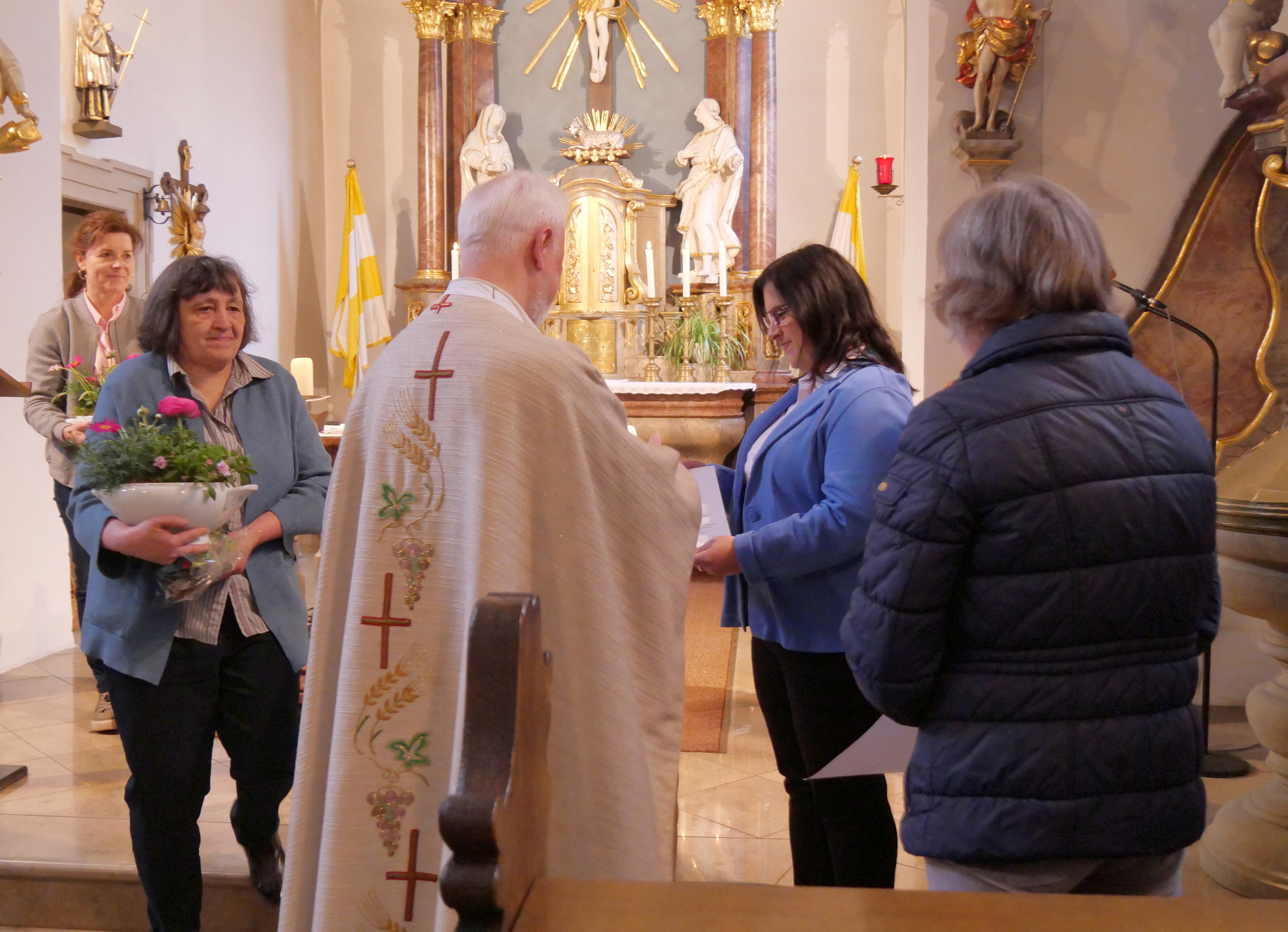 Das Bild zeigt eine kirchliche Zeremonie. Ein Priester in liturgischem Gewand steht vor einem Altar. Mehrere Frauen, eine mit Blumen, stehen vor ihm. Die Szene ist in einer Kirche mit Statuen und religiösen Symbolen.