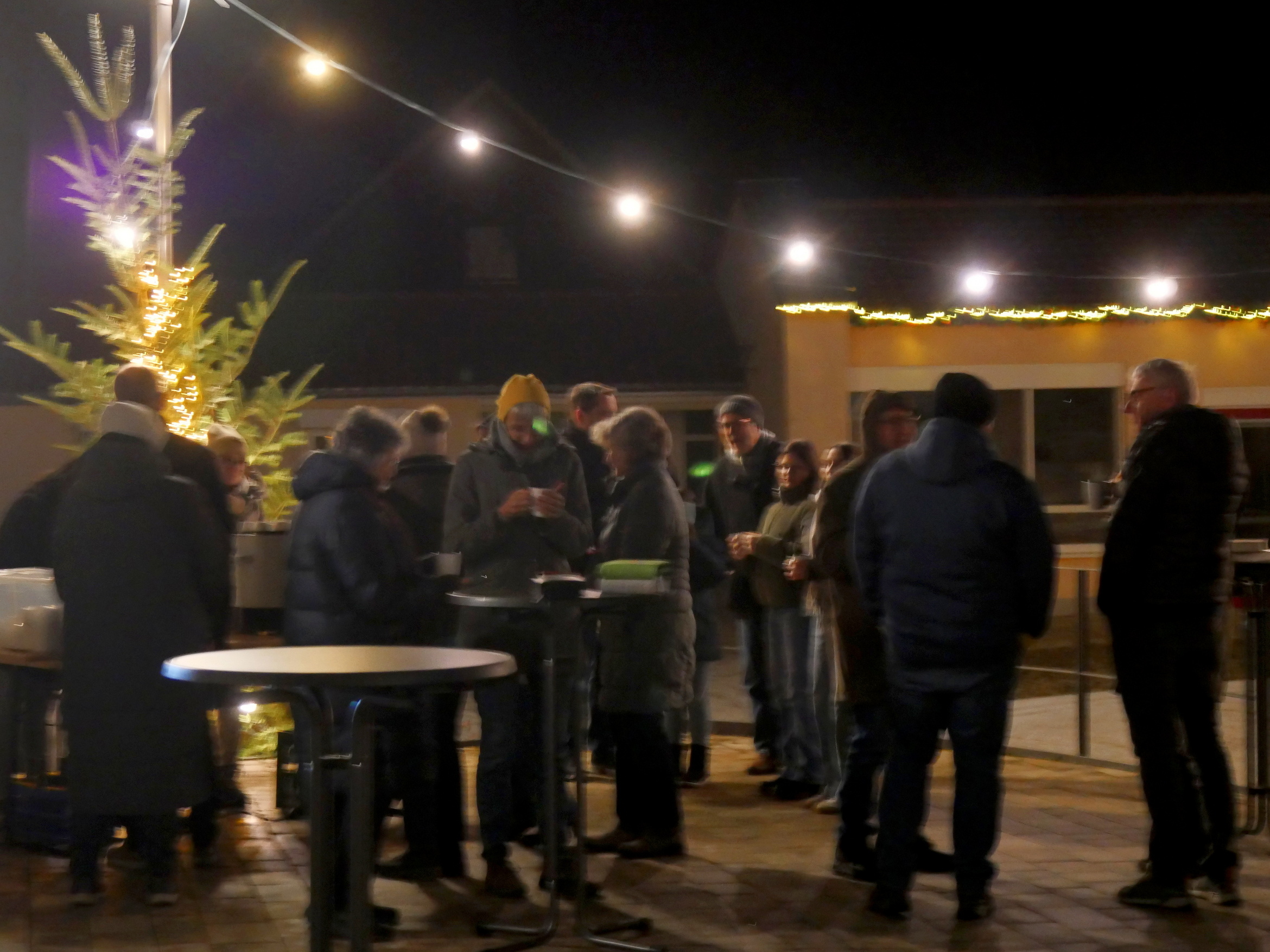 Auf dem Bild sind mehrere Personen auf dem Rathausplatz zu sehen. Es ist von einer gewissen Entfernung aufgenommen und etwas verschwommen. Man sieht, wie an Stehtischen Glüchwein getrunken wird.