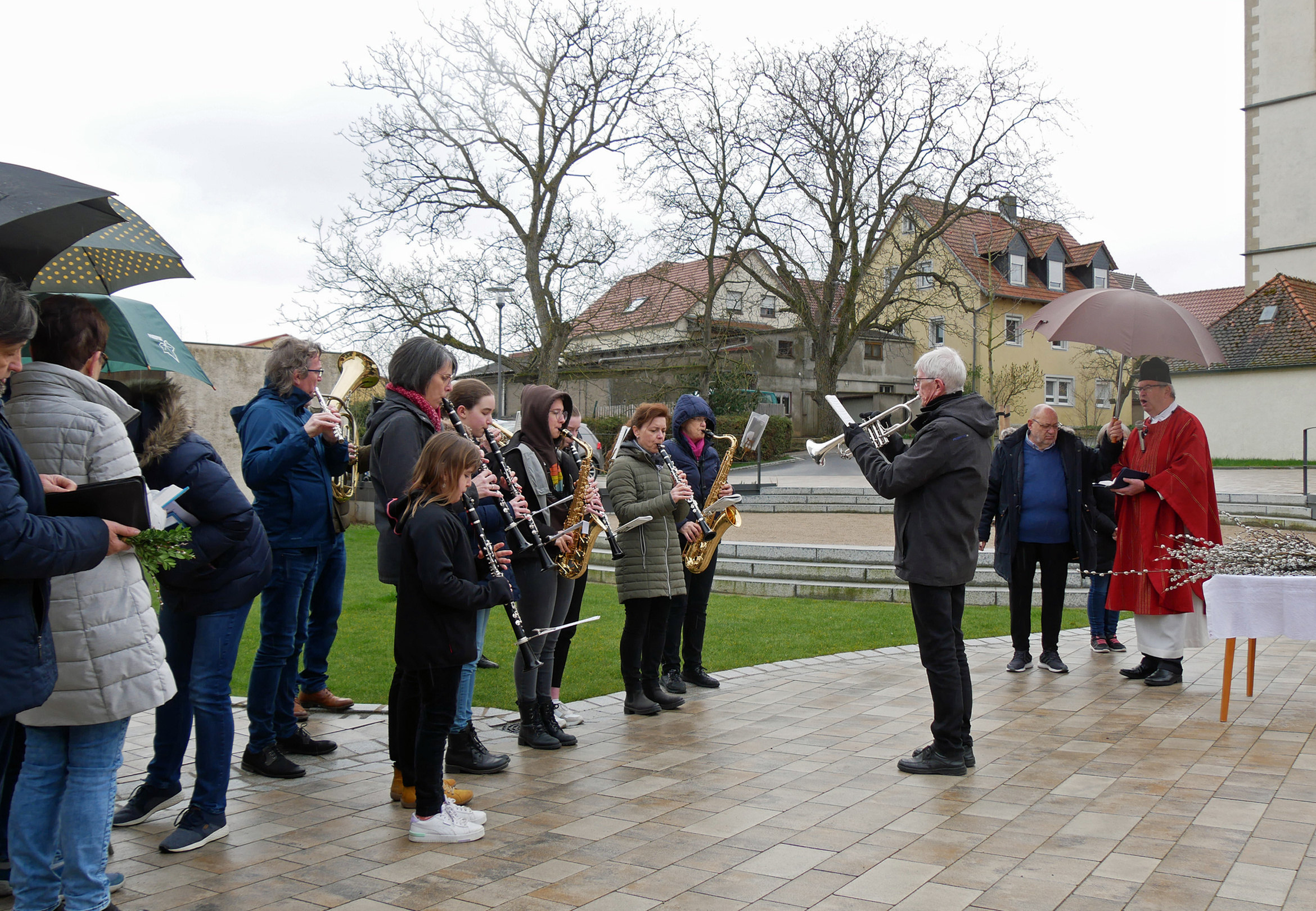 Die Musikkapelle spielt ein Lied. Rechts stehen der Pfarrer und der Küster. Es ist ein regnerischer Tag. Schirme sind aufgespannt.