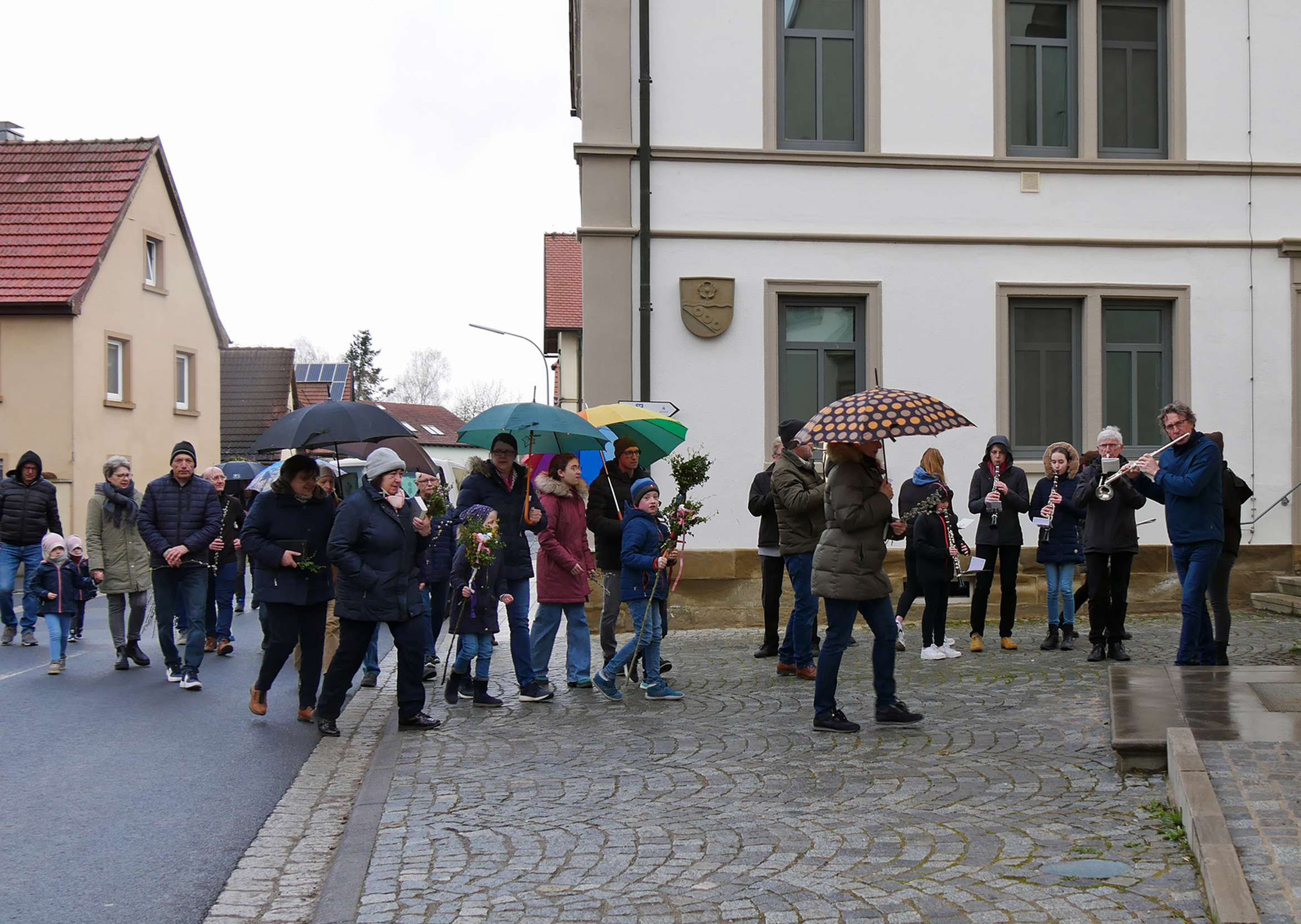 Mit einer Prozession ziehen Menschen in die Kirche ein. Es regnet, einige Regenschirme sind aufgespannt. Rechts steht eine Musikkapelle.