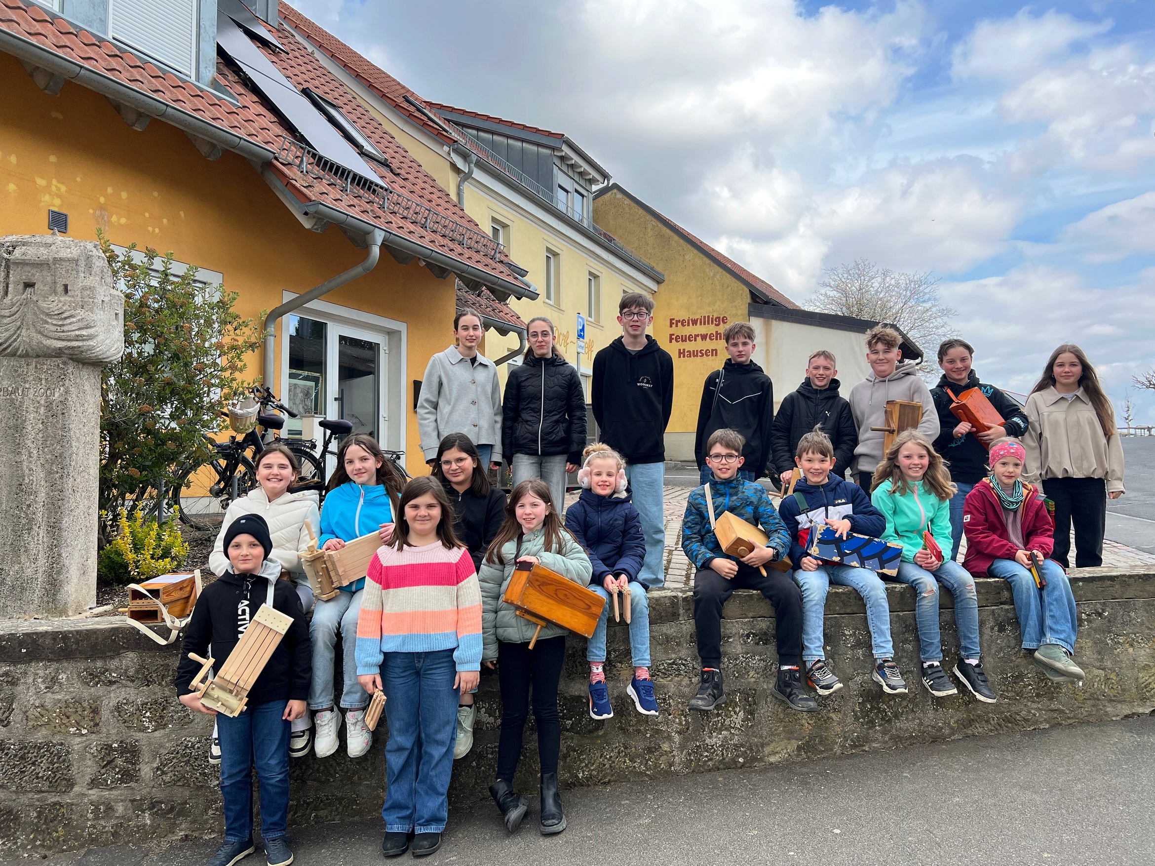 Das Bild zeigt eine Gruppe von etwa 20 Kindern und Jugendlichen, die vor einem gelben Gebäude mit der Aufschrift „Freiwillige Feuerwehr Hausen“ stehen. Einige sitzen auf einer Mauer, andere stehen. Viele halten selbstgebaute Holzobjekte. Die Stimmung ist fröhlich und entspannt.