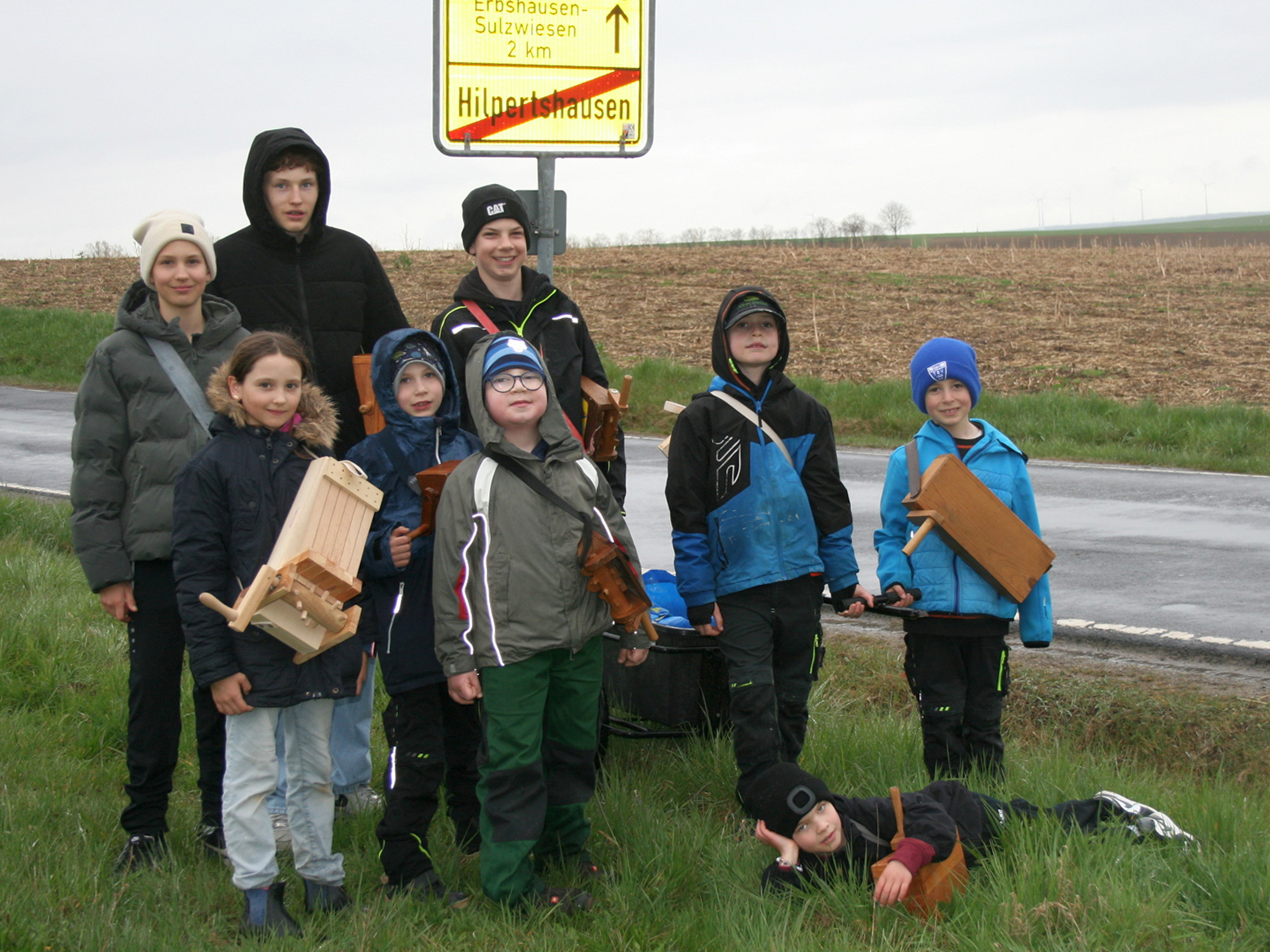 Das Bild zeigt eine Gruppe von neun Kindern, die an einer Straße stehen. Sie halten selbstgebaute Holzgegenstände und tragen wetterfeste Kleidung. Im Hintergrund ist ein Wegweiser mit Ortsnamen und Entfernungen zu sehen. Die Umgebung ist ländlich mit Feldern und einer Straße.