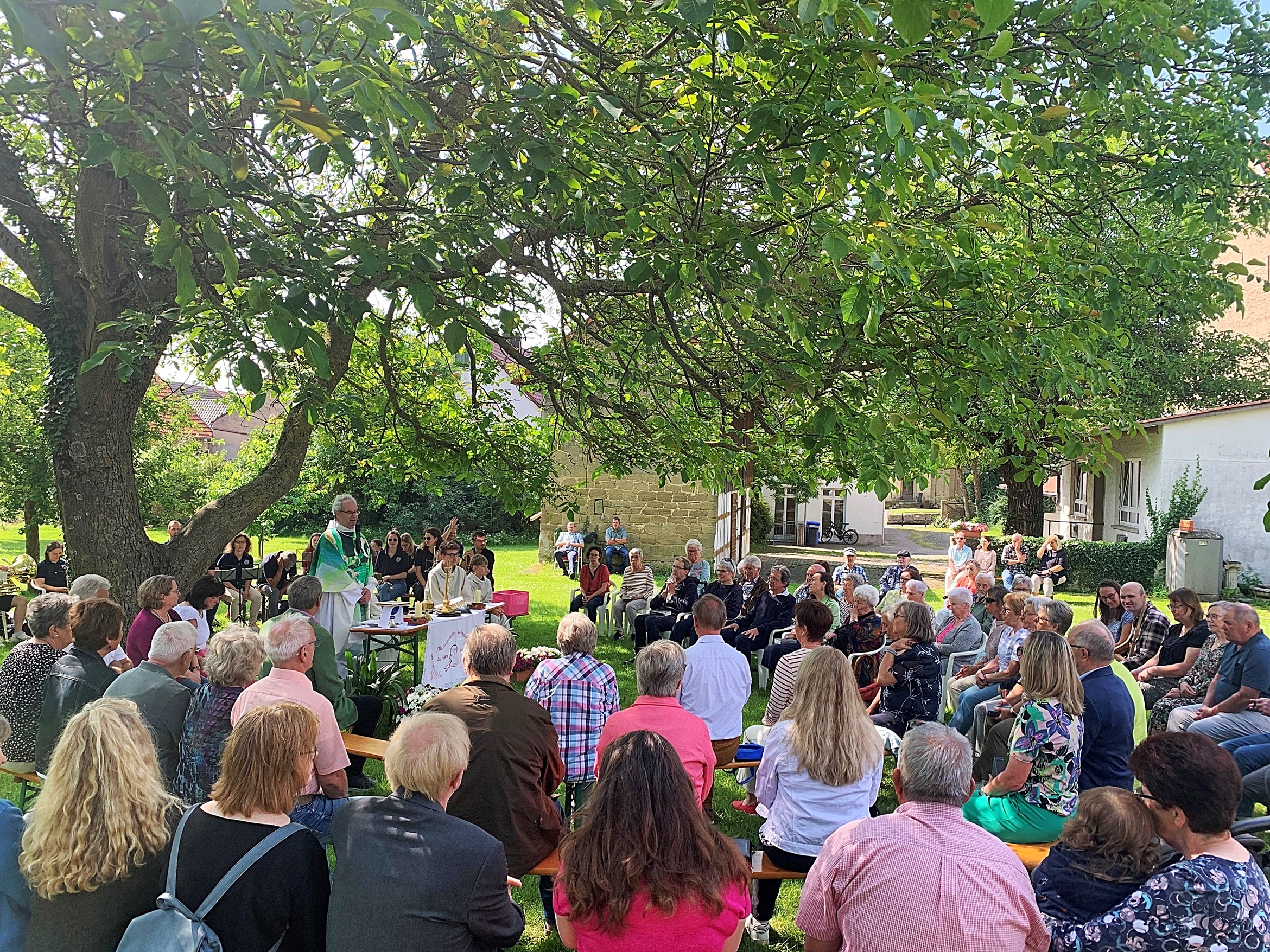 Unter einem großen Baum sitzen Menschen mit dem Rücken zur Kamera. Der Pfarrer steht hinten in der Mitte.