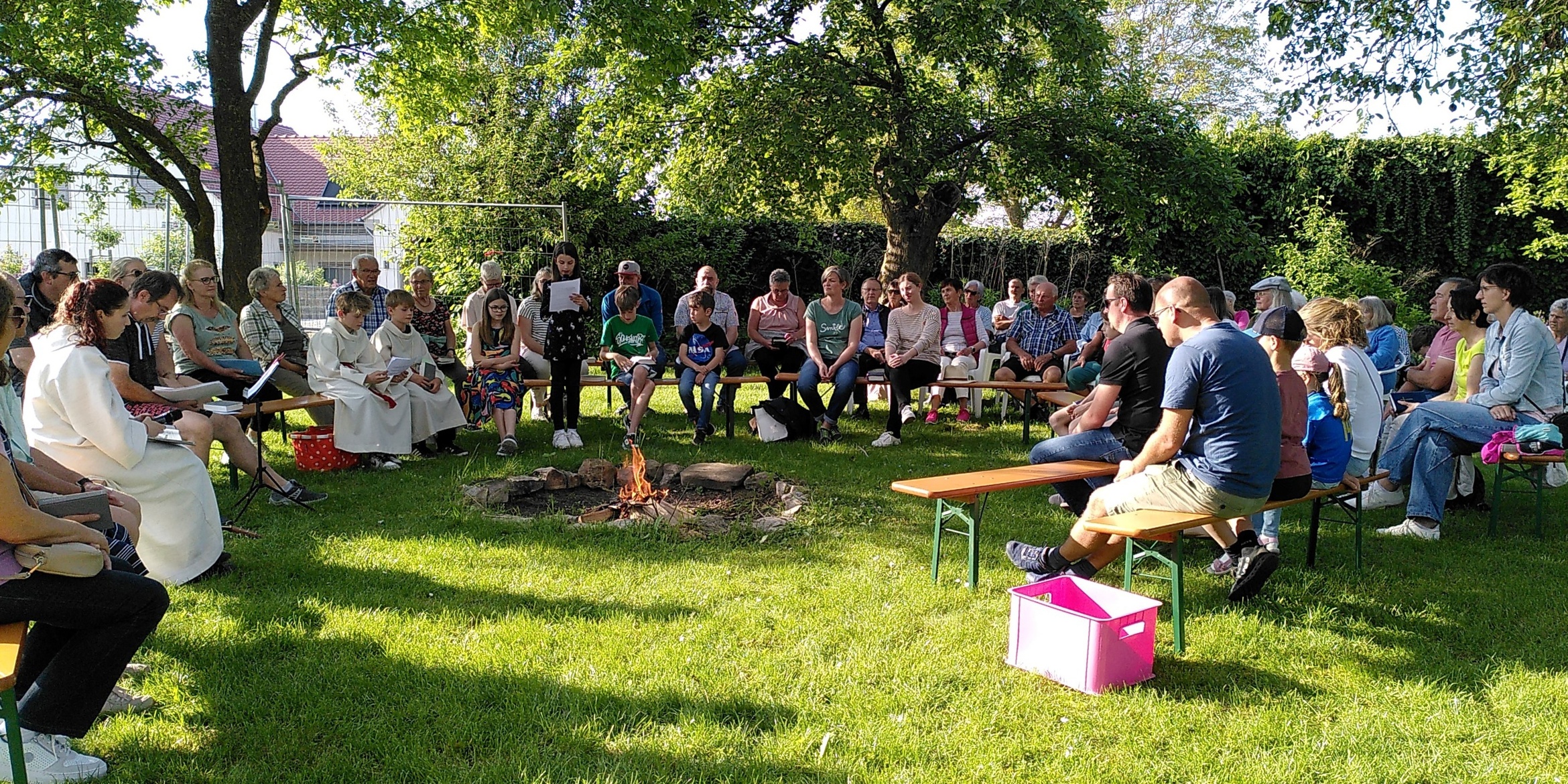 Im Grünen und im Kreisrund sitzen Menschen auf Bierbänken. Links sitzt Susanne Händel in einem weißen liturgischen Gewand.