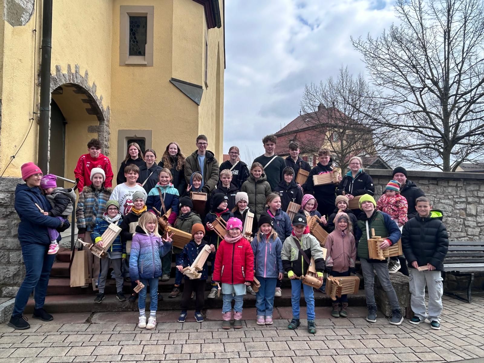 Etwa 35 Kinder und Jugendliche stehen auf einer Treppe und schauen in die Kamera. Sie halten Klappern aus Holz.