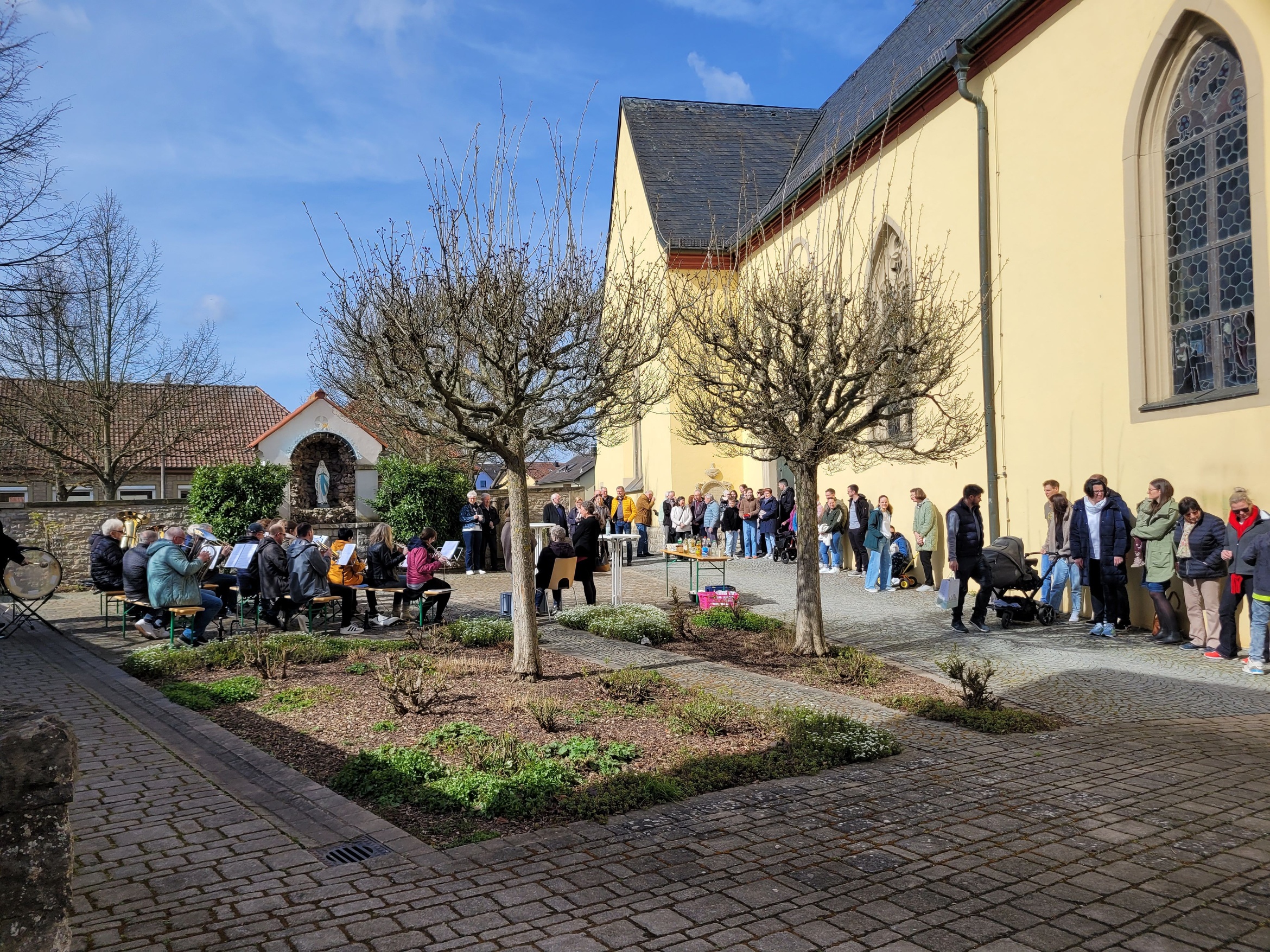 Blick auf den Kirchvorplatz, links sitzt die Blaskapelle und rechts an der Kirche stehen viele Menschen.