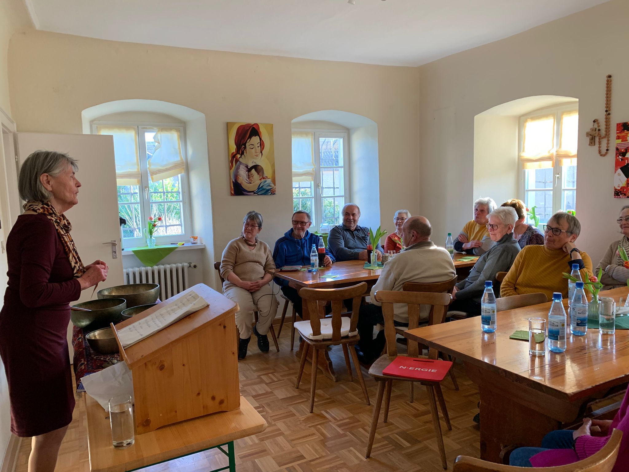 Links im Bild steht eine Frau an einem Tisch, auf dem mehrere Klangschalen stehen, und erzählt etwas. Rechts im Bild sieht man mehrere Leute, die an Tischen sitzen und aufmerksam zuhören.