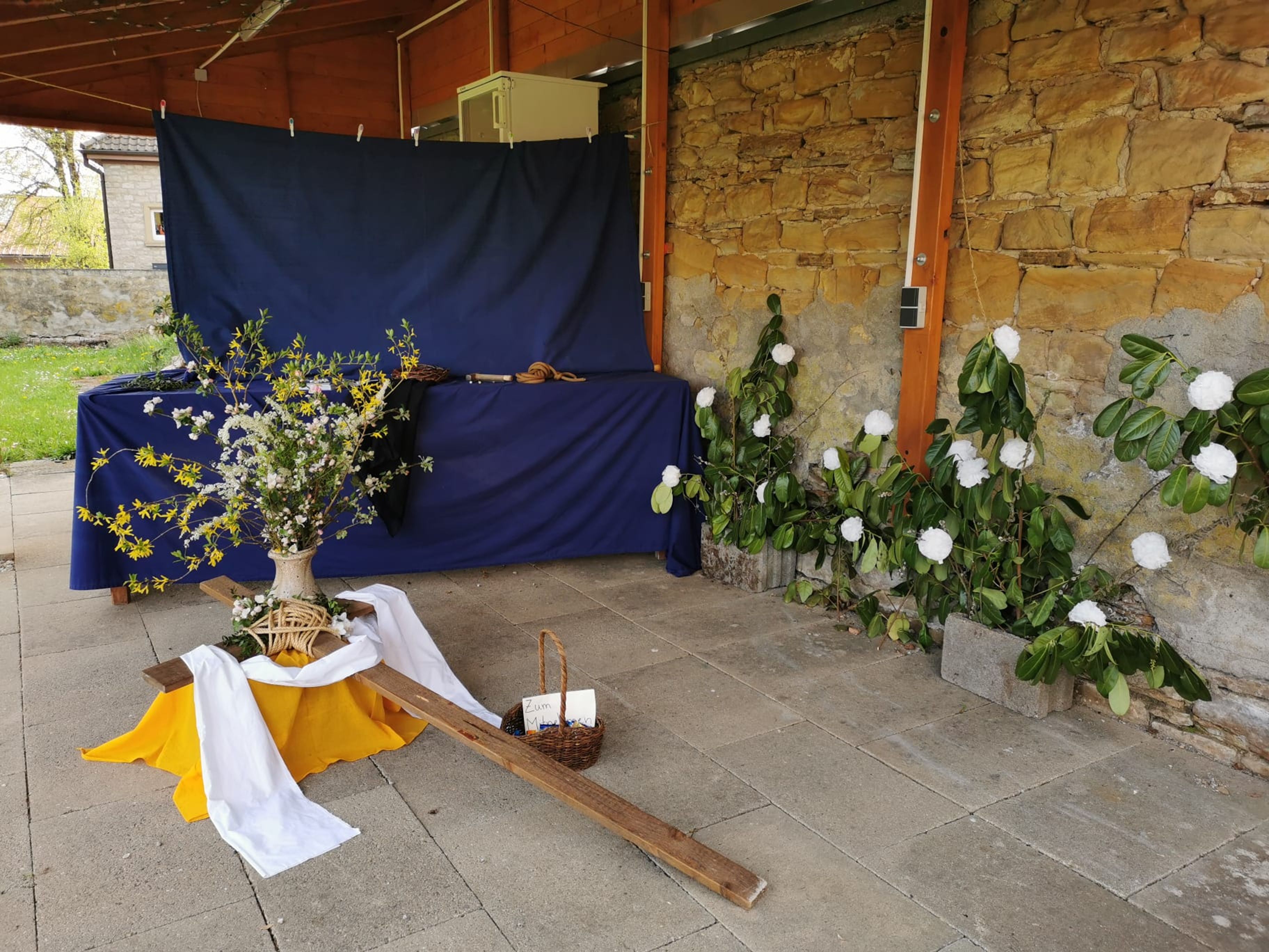 Im Vordergrund liegt ein Kreuz, es lehnt auf einem gelben Tuch, dahinter steht eine Blumenvase
