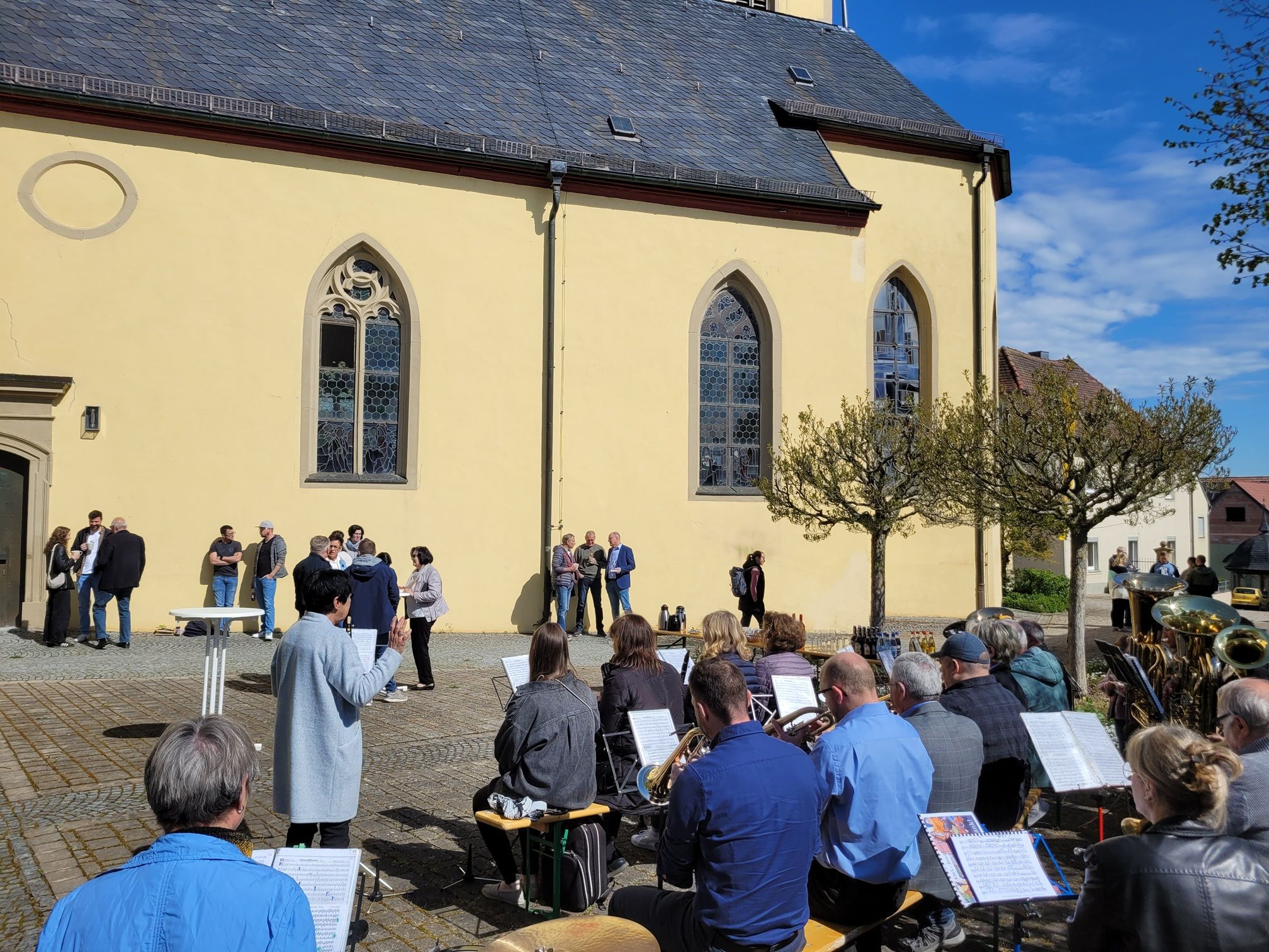 Blick von hinten auf die Blaskapelle, im Hintergrund stehen mehrere Menschen vor der Wand der Kirche und unterhalten sich.