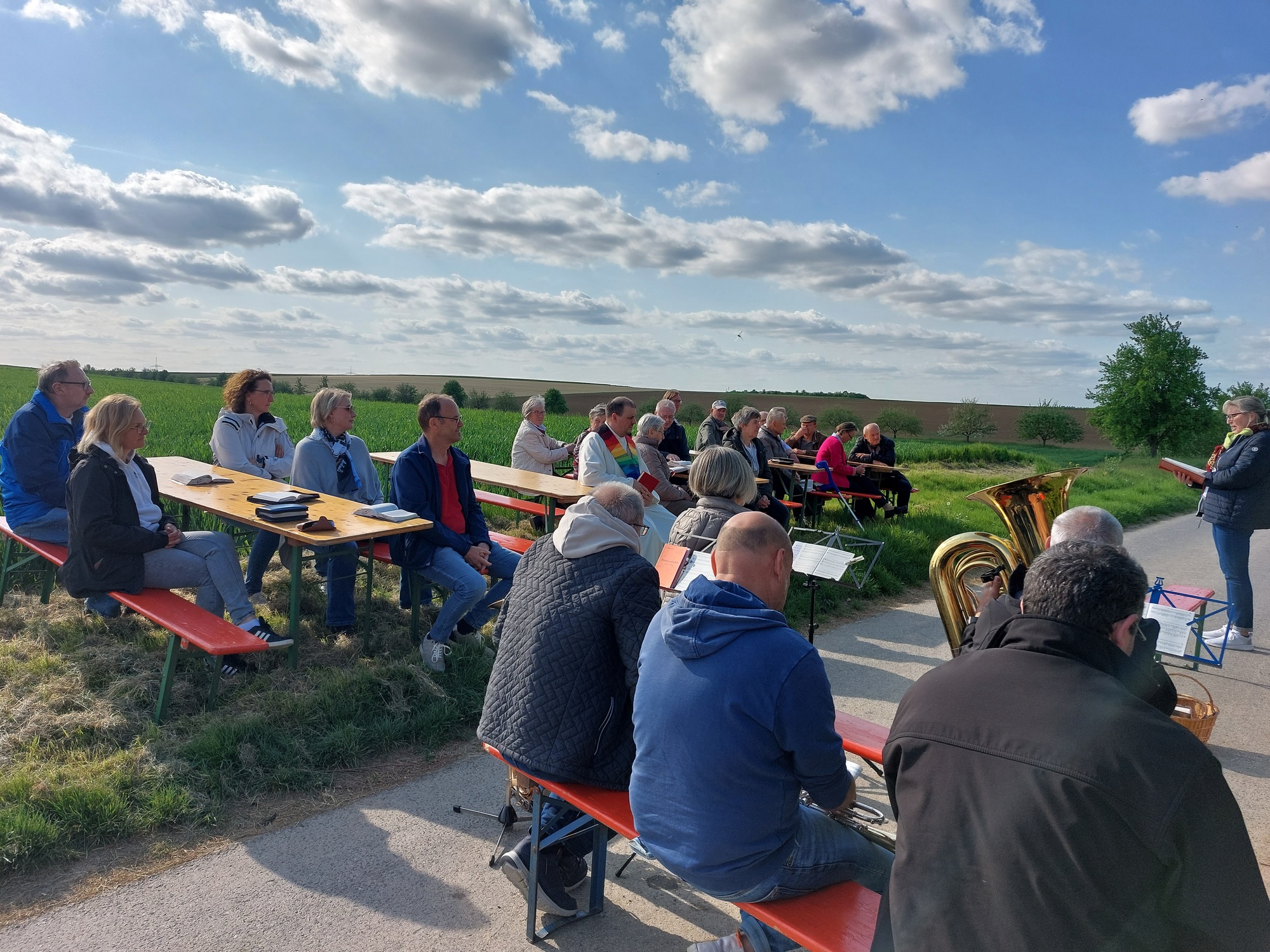 Unter weiß-blauem Himmel haben sich etwa 30 Personen versammelt zum Gottesdienst. Sie sitzen in Gruppen an Biertischen zusammen.