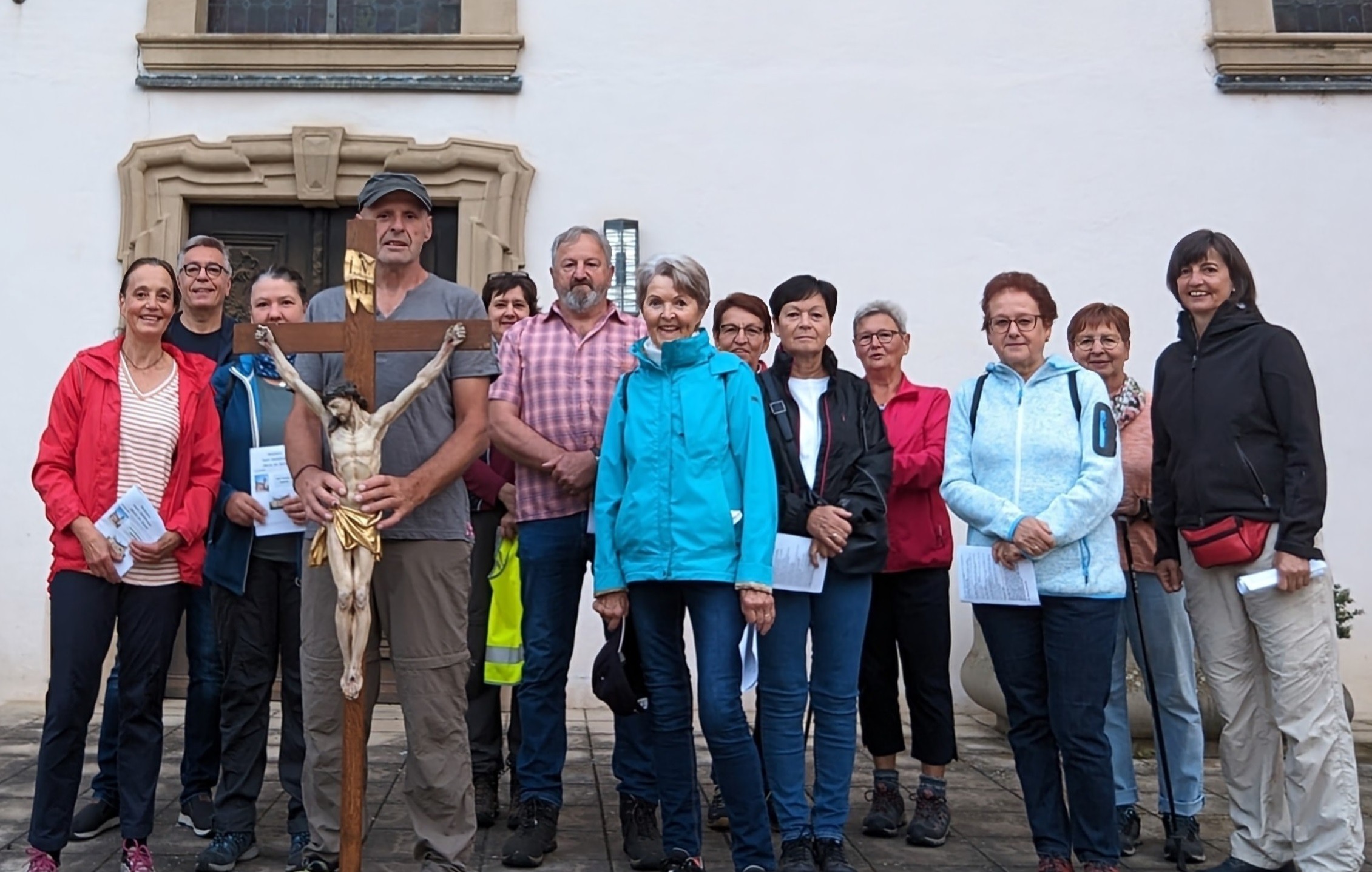 Eine Gruppe von 13 Menschen steht vor einer Kirche. Ein Mann hält vor sich ein großes Kreuz.