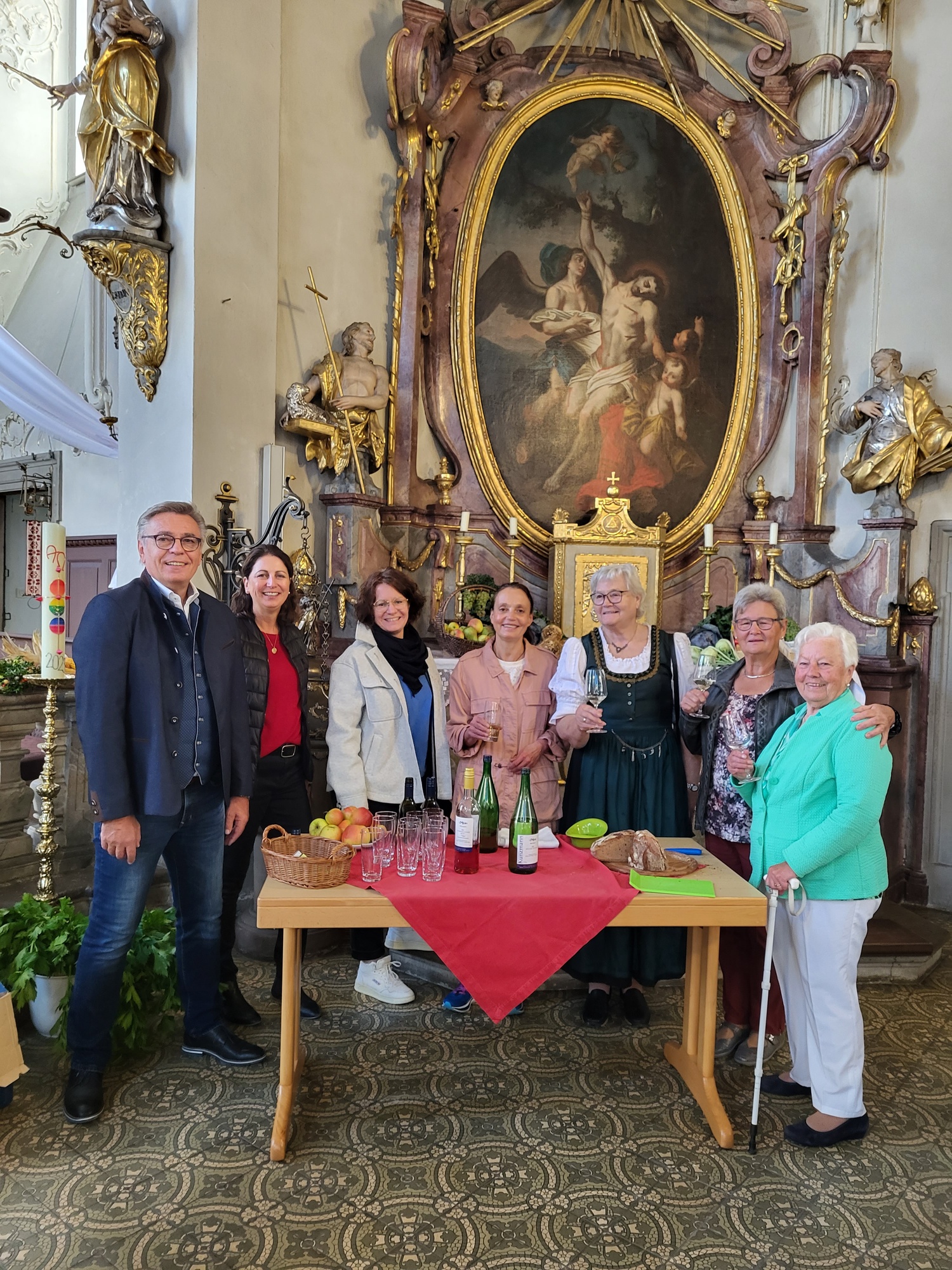 sieben Leute stehen in der Kirche um einen Tisch mit Erntegaben und Getränken
