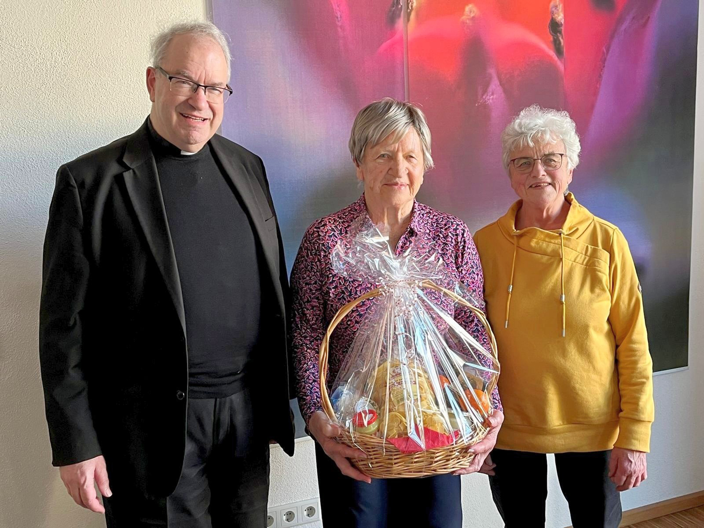 Auf dem Foto sind drei Personen. Sie schauen alle in die Kamera. Frau Brand in der Mitte hat einen Geschenkkorb in der Hand.