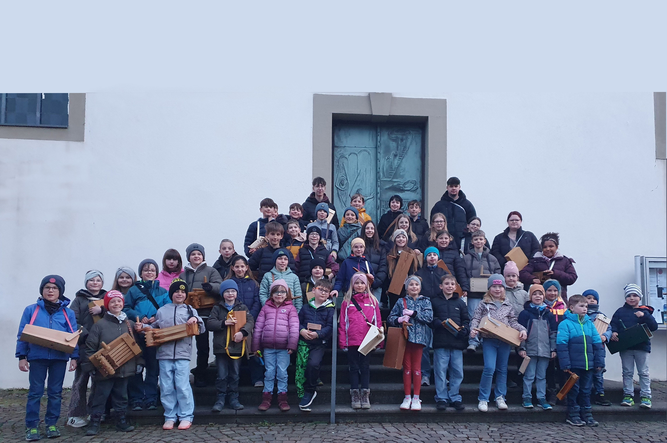 Das Bild zeigt eine Gruppe von etwa 50 Personen, darunter viele Kinder, die vor einem Gebäude mit einer großen Tür stehen. Sie halten hölzerne Kisten. Die Gruppe ist auf einer Treppe und einem Kopfsteinpflasterweg positioniert. Die Stimmung scheint fröhlich und organisiert zu sein.