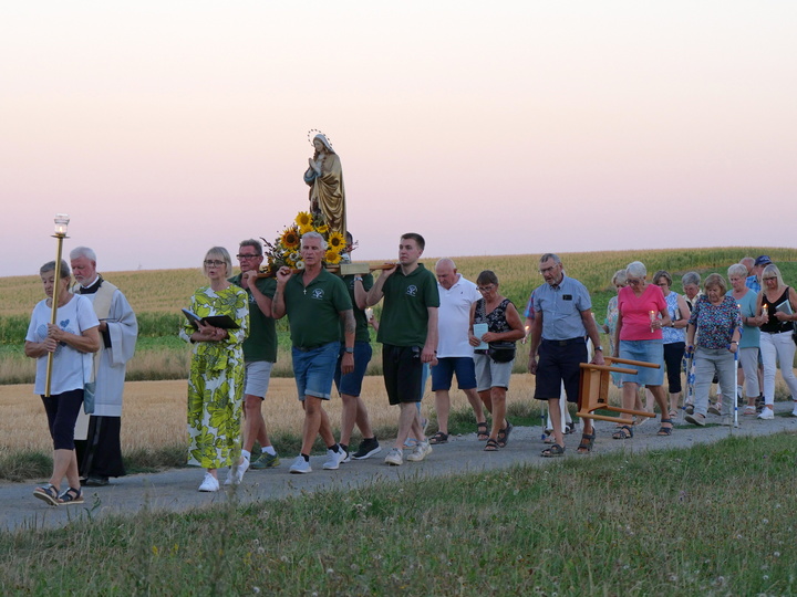Das Bild zeigt mehrere Menschen bei einer Prozession. Im Hintergrund sind Felder. In der Mitte ist die Marienstatue.