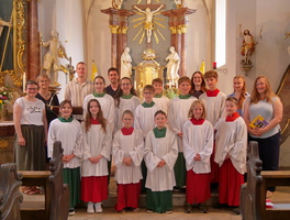 Auf dem Foto sind mehrere Kinder und Erwachsene in einer Kirche zu sehen. Die Kinder tragen weiße Chorhemden mit roten und grünen Schärpen, sie stehen vor einem Altar.