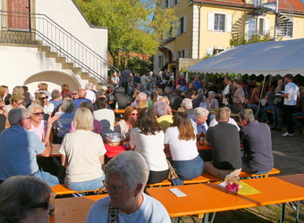 Das Bild zeigt einen Blick auf das Festgelände mit Tischen, Bänken und Ständen. Es ist sonniges Wetter und die Menschen unterhalten sich.