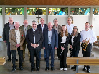 Auf dem Foto sind zehn Personen, darunter Musiker mit Klarinetten und einer Trompete, vor bunten Kirchenfenstern in einer Reihe stehend abgebildet. Sie lächeln.