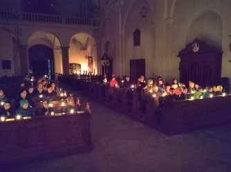 Das Bild zeigt eine dunkle Kirche, in der Menschen in Kirchenbänken sitzen und Kerzen in den Händen halten. Der Raum ist schwach beleuchtet, mit einem Altar im Hintergrund.