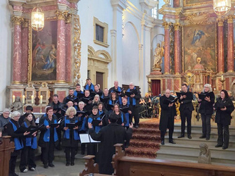 Das Bild zeigt einen Chor, der in einer Kirche singt. Die Mitglieder tragen schwarze Kleidung mit blauen Schals. Die Kirche hat prächtige, vergoldete Altäre und religiöse Gemälde im Hintergrund.