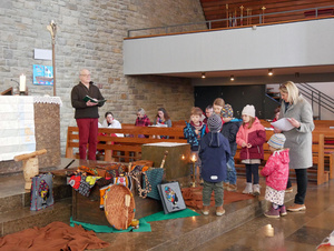 Im Altarraum der Kirche in Dipbach stehen zwei Erwachsene und zwischen ihnen mehrere Kinder. Es ist ein buntes Bild mit Stoffen und Gegenständen aus Mkoha auf dem Boden.