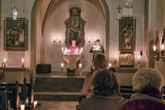 Der Pfarrer steht am Altar und breitet die Arme aus. Die Kirche ist dunkel. Kerzen brennen.