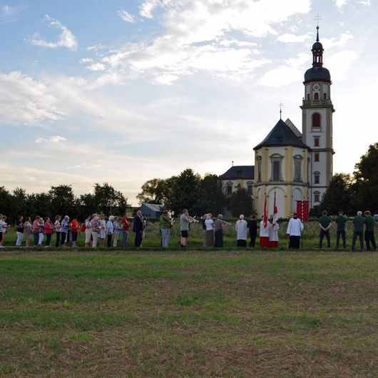 Das ist ein Blick über die Landschaft hinweg. Im Vordergrund ist ein Acker. Hinten sieht man klein die Wallfahrtskirche Fährbrück und davor eine Menschengruppe.
