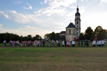 Das ist ein Blick über die Landschaft hinweg. Im Vordergrund ist ein Acker. Hinten sieht man klein die Wallfahrtskirche Fährbrück und davor eine Menschengruppe.