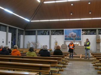 Das Bild zeigt das Innere einer modernen Kirche mit Holzbänken und einigen sitzenden Personen. Zwei Personen in orangefarbenen Warnwesten stehen vorne, eine mit einem Mikrofon. An den Wänden befinden sich Kunstwerke und Blumenarrangements.