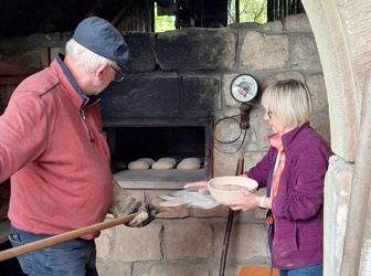 Das Bild zeigt einen Mann und eine Frau am Backofen. Sie kümmern sich um das Brotbacken. Die Brotlaibe um sie herum sind noch roh.