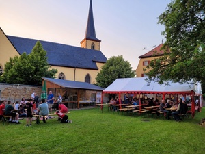 Blick auf den Pfarrgarten: Links mehrere Kinder am Lagerfeuer mit Stockbrot, rechts im Bild viele Erwachsene, die an Tischen sitzen und sich unterhalten.