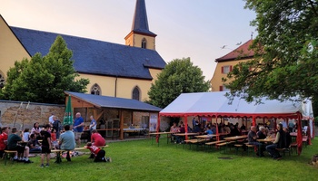 Blick auf den Pfarrgarten: Links mehrere Kinder am Lagerfeuer mit Stockbrot, rechts im Bild viele Erwachsene, die an Tischen sitzen und sich unterhalten.