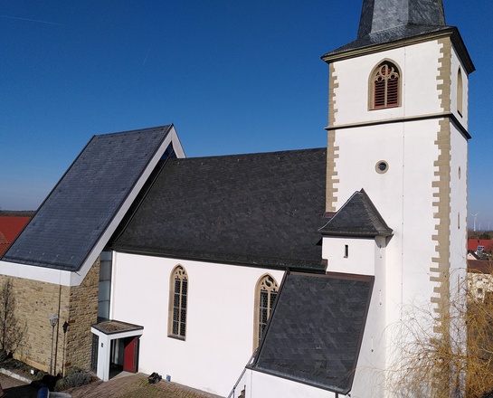 Das Bild zeigt eine weiße Kirche mit schwarzem Schieferdach, spitzem Turm und gotischen Fenstern unter klarem blauem Himmel in einer ländlichen Umgebung.