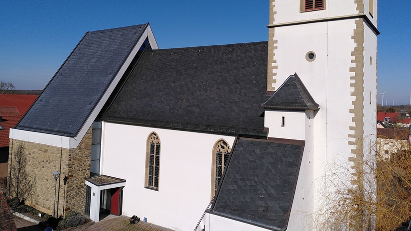 Das Bild zeigt eine weiße Kirche mit schwarzem Schieferdach, spitzem Turm und gotischen Fenstern unter klarem blauem Himmel in einer ländlichen Umgebung.