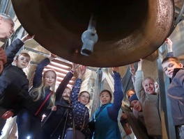 Ein Gruppe von Kindern steht unter einer großen Glocke in einem Glockenturm. Die Kinder sind verschieden gekleidet, manche tragen Mützen. Die Glocke befindet sich über ihnen und die Kinder schauen nach oben.